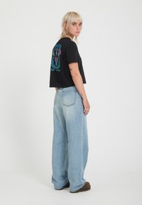 Jeune personne aux cheveux blonds courts porte un t-shirt graphique noir et un jean large bleu clair, debout de profil sur un fond blanc uni.