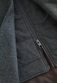 Tessuto ad herringbone grigio con superficie texturizzata, dotato di una cerniera centrale e di una fodera liscia. L'interno presenta un colore marrone solido.