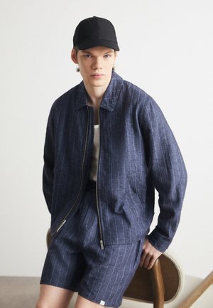 Veste rayée bleu foncé avec une fermeture éclair et des manches courtes, assortie à un short. Casquette noire, assise sur une chaise ronde contre un fond neutre.