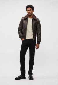 Καφέ δερμάτινο μπουφάν με γούνα στον γιακά, μπεζ πουλόβερ από κάτω, μαύρο παντελόνι και μαύρα παπούτσια. Λείο υλικό και κομψή γραμμή.