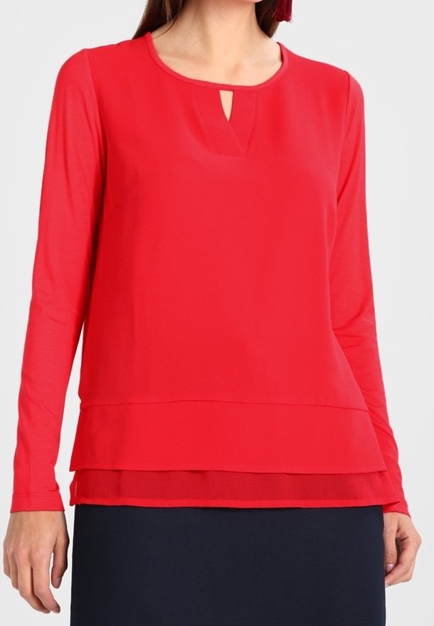 Woman wearing a bright red long-sleeve blouse with a layered hem and a small keyhole neckline, paired with a dark skirt.