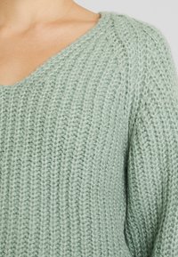 Ljusgrön stickad tröja med V-ringning, prydd med ett texturerat ribbmönster. Mjuk, tjock garn ger ett mysigt utseende. Inga synliga detaljer.