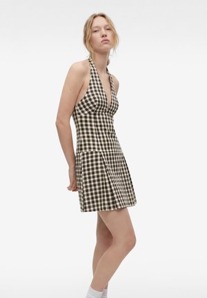 Jeune femme portant une robe à carreaux noire et blanche sans manches, debout les mains derrière le dos sur un fond blanc.