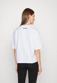 Valkoinen puuvillapaita, jossa on lyhyt malli; siinä on hieman laskeutuvat hartiat ja musta "KARL LAGERFELD" -logo takana kaula-aukon kohdalla.