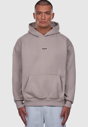 Mężczyzna w beżowej bluzie z kapturem i kieszenią z przodu, z logo "Dropsize" na środku klatki piersiowej, w jasnoniebieskich dżinsach, stojący na jednolitym tle.