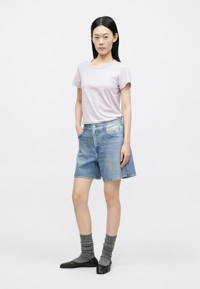 Jovem mulher está de pé, vestindo uma t-shirt lilás clara, calções de ganga de cintura alta, meias cinza franzidas e sapatilhas pretas, sobre um fundo branco.