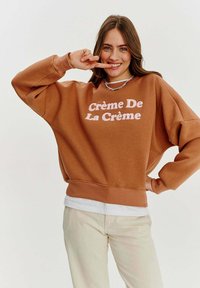 Hnědý oversized svetr s růžovým výšivkou "Crème De La Crème", s žebrovými manžetami a lemem. Nositý přes bílou košili, v kombinaci se světlými kalhotami.
