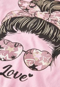 Grafisk t-shirt med en pink baggrund, stiliseret hår med et mønstret sløjfe og hjerteformede solbriller med stjernemønstre. Ordet "Kærlighed" er i sort glimmer.