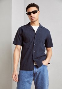 Camisa de manga corta de tejido texturizado en azul marino con botón en el frente, un solo bolsillo en el pecho y corte recto, combinada con unos vaqueros de mezclilla azul claro.