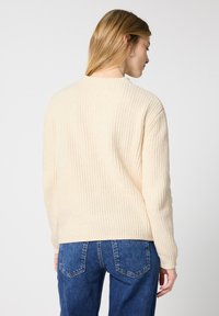 Maglione a costine crema con maniche lunghe, scollatura rotonda e vestibilità rilassata, abbinato a jeans in denim blu. Tessuto testurizzato con una finitura morbida.