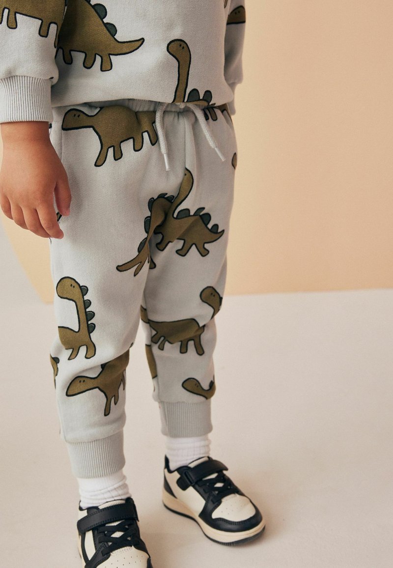 Grå sweatpants med dinosaurtryk i grøn og sort. Elastisk talje med snor, ribstrikkede manchetter, parret med sort og hvidt sneakers.