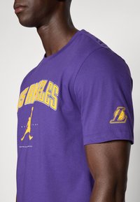Violeta kokvilnas T-kreklu ar uzrakstu "LOS ANGELES" dzeltenā krāsā, basketbola siluetu un Lakers logo uz piedurknes. Ar īsām piedurknēm.