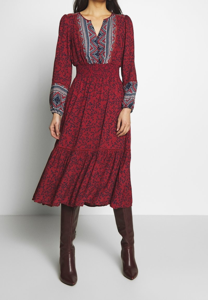 Femme portant une robe midi rouge fleurie avec des manches longues à motifs et un buste à motifs, associée à des bottes hautes marron foncé, debout contre un mur blanc.