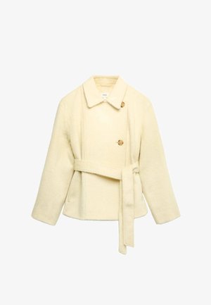 Giacca color crema con tessuto morbido e strutturato, caratterizzata da un colletto a revers, doppia fila di bottoni e cintura annodata in vita.