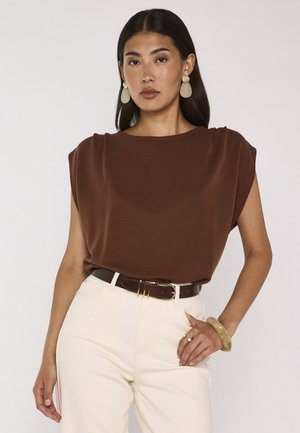 Femme aux longs cheveux foncés porte un haut marron sans manches, un pantalon crème taille haute, une ceinture foncée, de grandes boucles d'oreilles crème et un bracelet assorti.