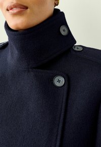 Manteau en laine marine avec col montant, présentant un design croisé, deux grands boutons et une finition texturée.