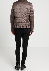 Brązowa pikowana kurtka z falbaniastym dołem, gładką teksturą i kołnierzem. W połączeniu z czarnymi dopasowanymi spodniami i botkami za kostkę.