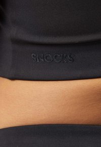 Crop top nero realizzato in un tessuto morbido con una vestibilità aderente; presenta il logo "SNOCKS" in rilievo lungo l'orlo. Area color pelle visibile sopra il top.