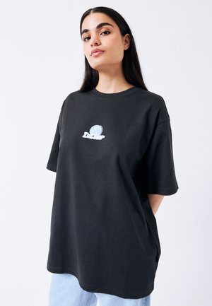 Jonge vrouw met lang donker haar die een oversized zwart T-shirt draagt met een klein rond blauw-wit logo op de borst.