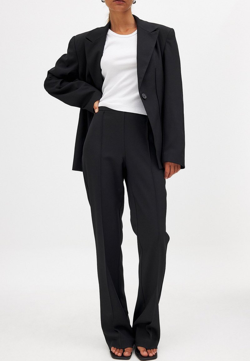 Črn blejzer s strukturiranimi ramenimi deli, v kombinaciji z visokimi črnimi hlačami. Pod njim je bela prilegajoča se rebrasta majica. Gladka tekstura, natančna postava.