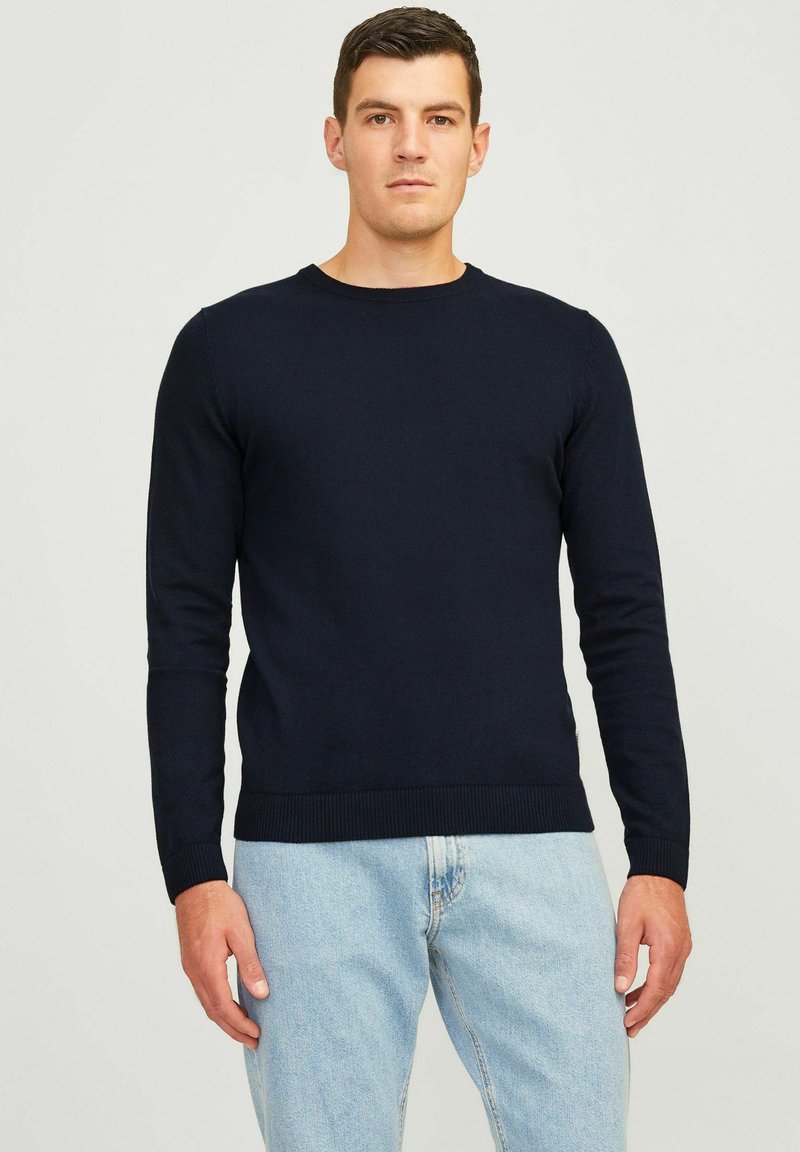 Homme portant un pull noir uni à manches longues et un jean bleu clair, debout devant un fond neutre.