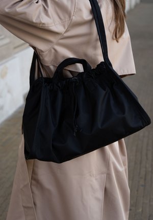 Kvinde i beige trenchcoat bærer en stor sort snøresæk over skulderen mod en udendørs baggrund.