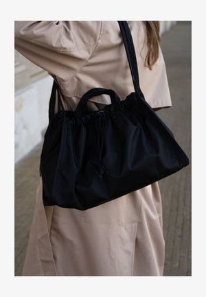 Femme en trench beige portant un grand sac à main noir à cordon sur l'épaule, sur un fond extérieur.