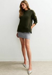 Maglione oversized verde oliva in maglia testurizzata, abbinato a una mini gonna grigia. Calzature: décolleté argentate a punta con decorazioni.