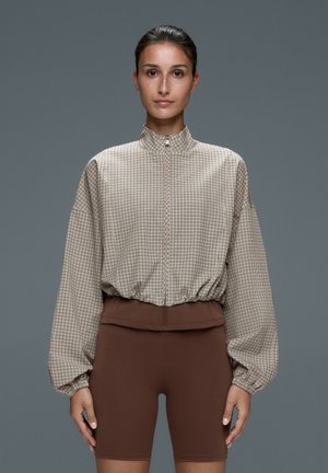 Frau, die nach vorne schaut, trägt eine beige karierte Reißverschlussjacke mit Ballonärmeln und braune eng anliegende Shorts.