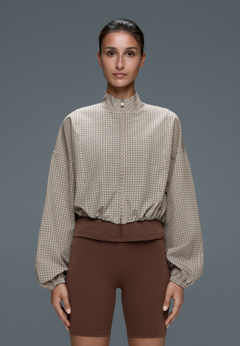 Femme debout face à nous portant une veste beige à carreaux zippée avec manches ballon et shorts marron ajustés.