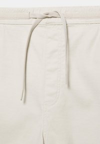 Pantalon beige clair avec un cordon en tissu noué à la taille et des coutures visibles le long de l'ouverture avant.
