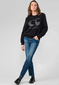 Sweatshirt noir avec un logo argenté, assorti à un jean bleu déchiré et des chaussures noires. Le tissu semble doux, avec une coupe décontractée.