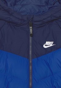 Námořnická péřová bunda s modrými panely, se zapínáním na zip a malým bílým logem Nike na hrudi. Hladký, texturovaný materiál.
