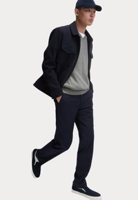 Giacca blu con due tasche sul petto, maglione grigio, pantaloni blu a righe e sneakers blu con suole bianche. Il modello indossa un cappellino abbinato.