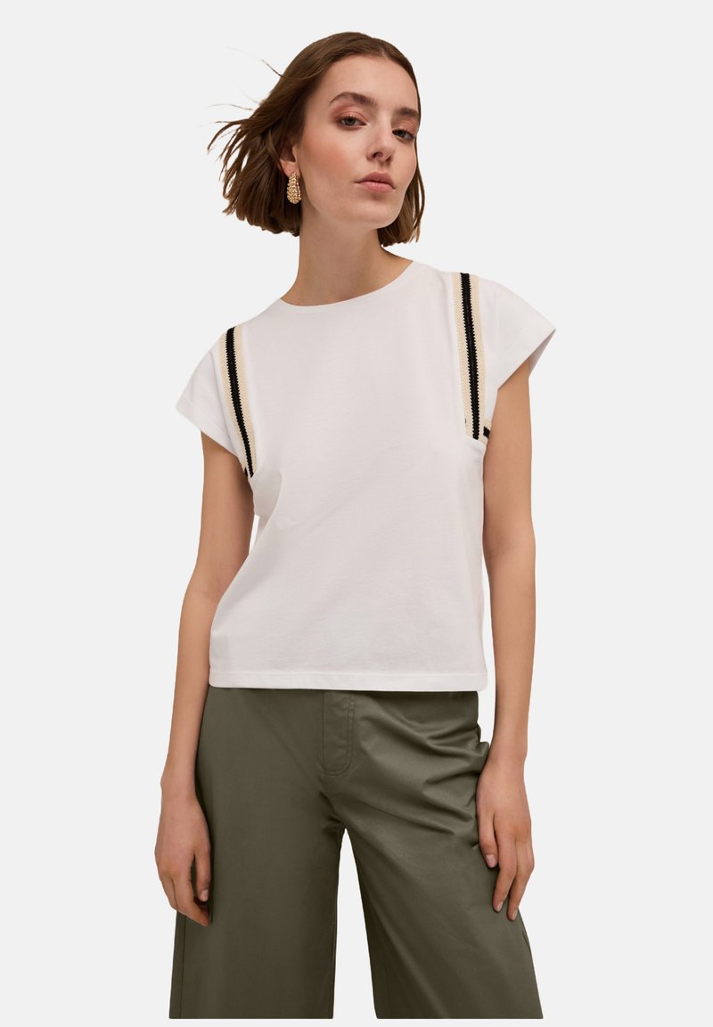 Tricou alb din bumbac cu mâneci scurte, având dungi negre și crem pe umeri. Asortat cu pantaloni largi de culoare verde măslin.