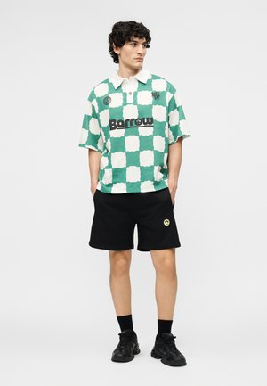 Unge mann iført grønn- og hvitrutete skjorte med "Barrow"-logo, svarte shorts med gult smilemerke, svarte sokker og svarte sneakers.