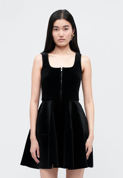 Femme aux longs cheveux noirs portant une robe en velours noire sans manches avec une fermeture éclair sur le devant et des boucles d’oreilles dorées sur un fond blanc uni.