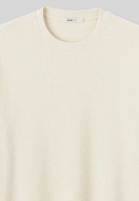 Pull à manches longues en maille côtelée de couleur écrue, avec un col rond et un tissu texturé. Conçu pour offrir une coupe décontractée avec des détails de couture subtils.