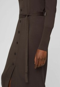 Brązowa dzianinowa sukienka z guzikiem z przodu, długimi rękawami i paskiem w talii z bocznym rozcięciem dla dodatkowego akcentu.