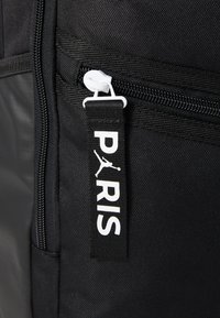 Fekete hátizsák cipzáras zsebbel, fehér cipzárhúzóval, valamint egy textil címkével, amelyen „PARIS” és egy logó látható kontrasztos színekben.