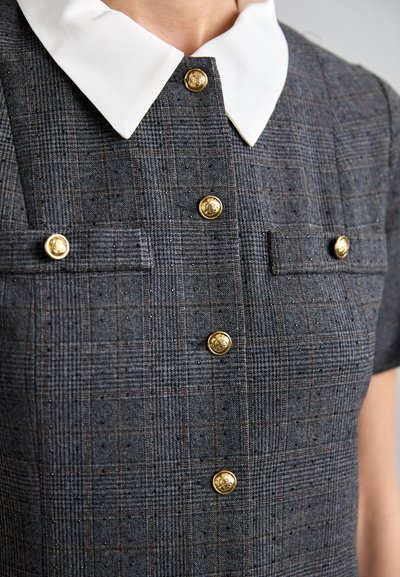 Rochie gri în carouri, cu o cămașă albă, butoni aurii și două buzunare pe piept, având o textură fină și un design structurat.
