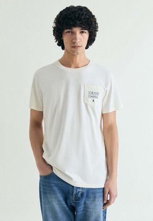 Junger Mann mit lockigem Haar, der ein weißes "Scalpers Company"-T-Shirt und blaue Jeans trägt und vor einem schlichten hellen Hintergrund steht.