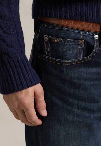 Prikaz ruke pokraj tamnoplave Polo džepne traperice s narančastim šavom, nošene s smeđim remenom i tamnoplavo pletenim džemperom.