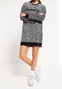 Graues Strickkleid mit schwarzen Akzenten, das einen Logo-Druck und seitliche Reißverschlussdetails aufweist. Kombiniert mit weißen Sneakers und gerippten Bündchen.