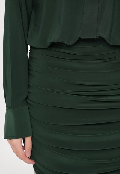 Tmavozelená blúzka skombinovaná s riasenou sukňou. Materiál má hladkú textúru a rukávy sú zakončené zvonovitými manžetami.