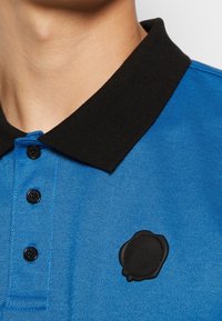 Gros plan sur une chemise polo bleue avec un col et des boutons noirs, ornée d'un logo en style sceau noir sur la poitrine.