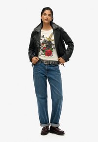 Veste en cuir noir de moto sur un T-shirt graphique avec un design de loup et de rose, jean bleu, et mocassins marron avec une semelle épaisse.