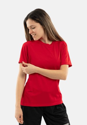 Jonge vrouw die lacht, draagt een eenvoudige rode korte mouw t-shirt en zwarte broek, met linkerhand zorgt ze dat de mouw goed zit, witte achtergrond.
