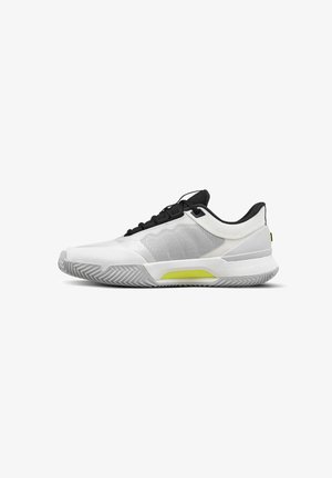 Zapatilla deportiva blanca de corte bajo con cordones negros, panel lateral de malla, suela acolchada y detalle amarillo en la entresuela.