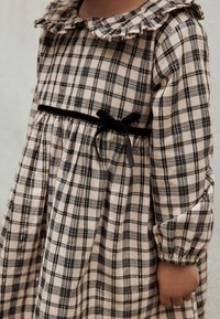 Plaid dress in beige and dark colors, featuring a ruffled collar, velvet waistband, and gathered sleeves for texture and detail.